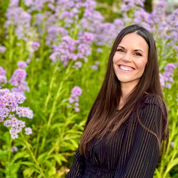Smiling woman in black blouse posing outdoors with purple flowers.
