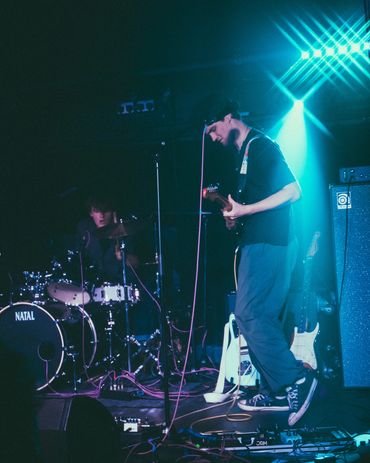 Young guitarist and drummer performing live on stage with blue lighting.