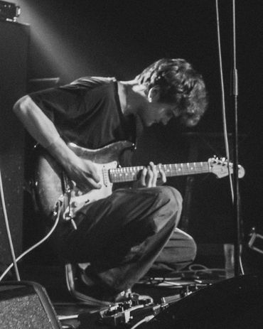 Musician intensely playing electric guitar on stage, surrounded by equipment and spotlights.