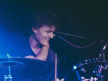 Drummer smiling while playing a Natal drum set on stage with blue lighting.