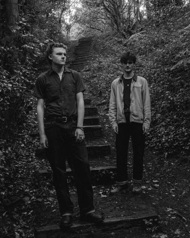 Two young men standing on a forest trail with steps, surrounded by dense trees.