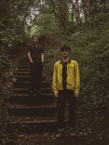 Two young men stand on a forest path with stone steps surrounded by dense greenery.