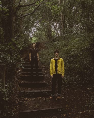 Two young men standing on forest stairs surrounded by dense greenery.