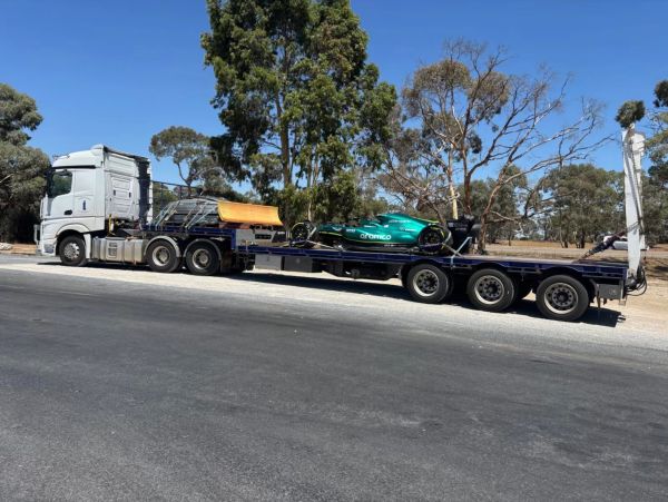 A truck carrying a green race car on its flatbed trailer on a sunny day.