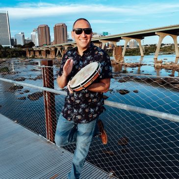 Man playing a tambourine by the river, smiling with sunglasses.