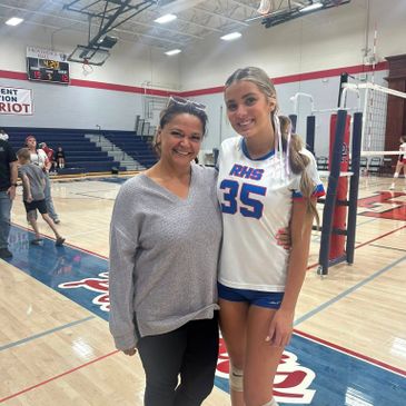 A volleyball player in uniform poses with a smiling woman in a gymnasium.