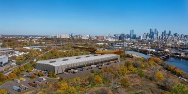 A large industrial warehouse near a river with a city skyline in the background.