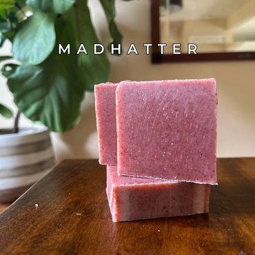 Three pink square soap bars stacked on a wooden surface with a plant in the background.