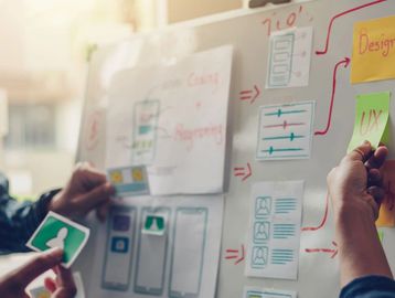 Hands place sticky notes on a wall showing wireframes, user flows, and UX design ideas.