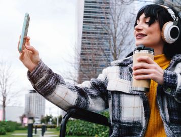 Person wearing headphones holds a phone and coffee while sitting outdoors, smiling at the screen.