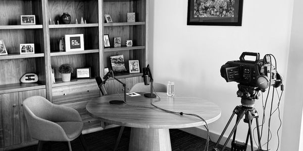 A podcast setup with two microphones at a round table, a camera on a tripod, and shelving in back.