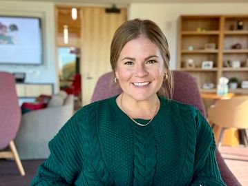 Smiling woman in a green sweater sits indoors in a warm, modern workspace.