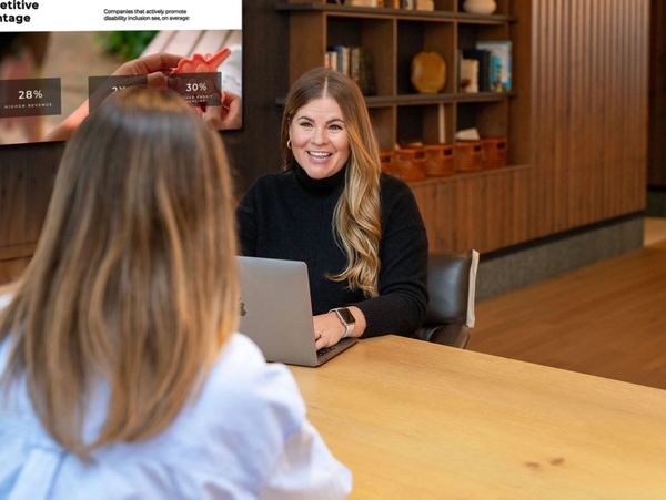 Woman smiling during a meeting, seated at a table with a laptop, speaking with another person.