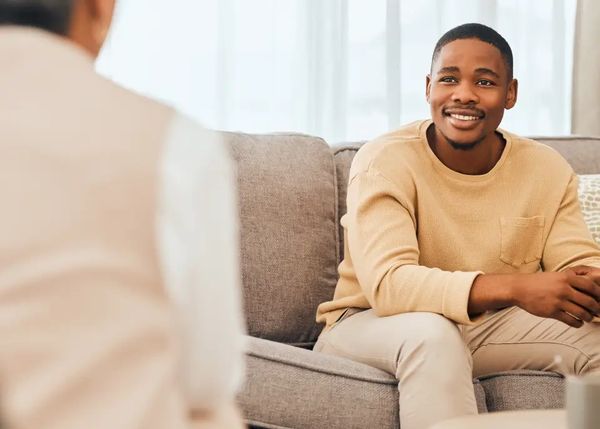 A smiling Black man in a beige sweater and light pants, seated on a gray sofa, looking right