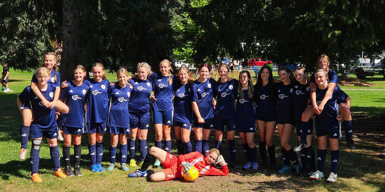 A girls' soccer team posing outdoors in blue jerseys with their goalie lying in front holding a yellow ball.