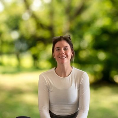 Smiling woman in white shirt sitting outdoors with blurred green background.