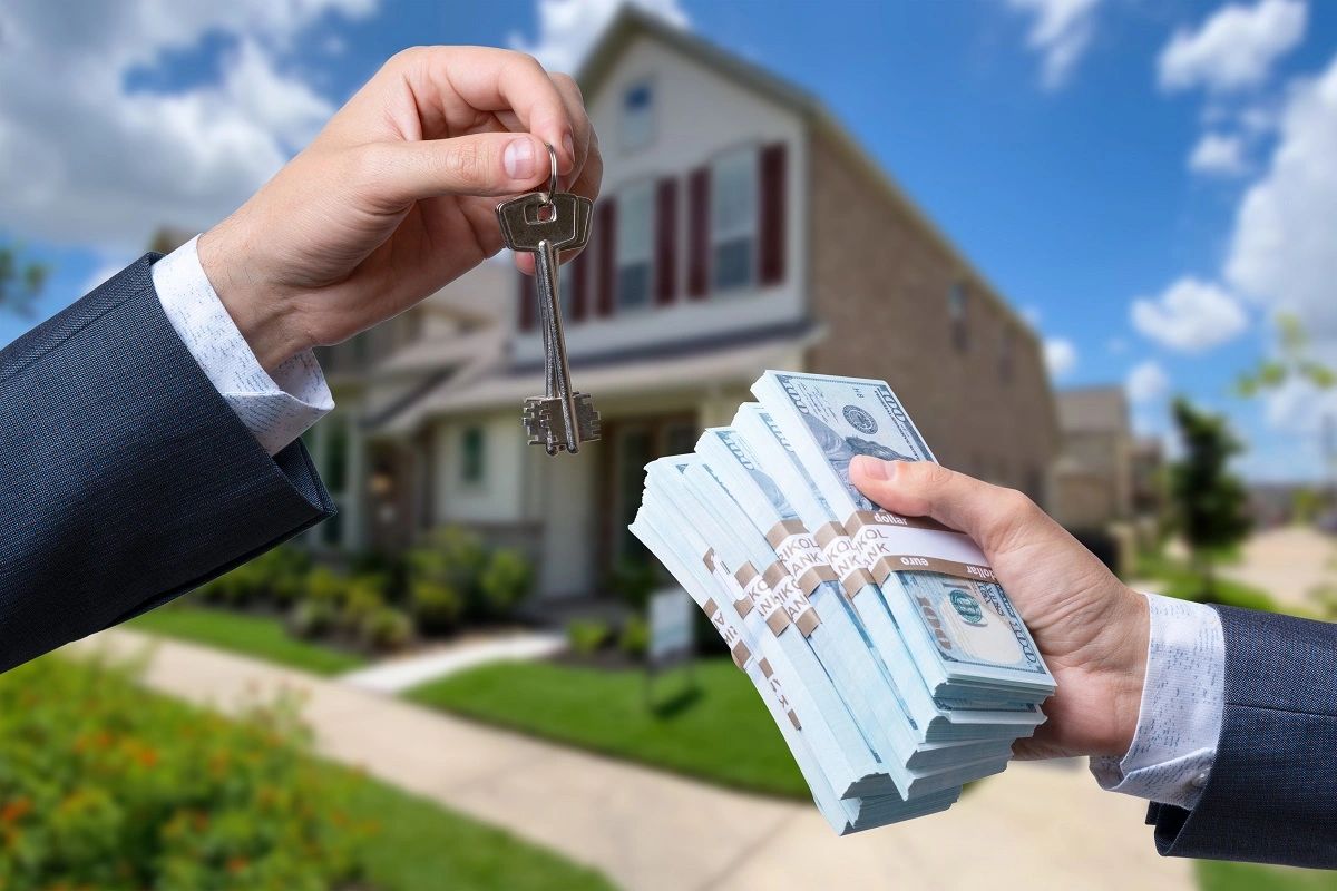 Hand exchanging house keys for stacks of cash in front of a house.