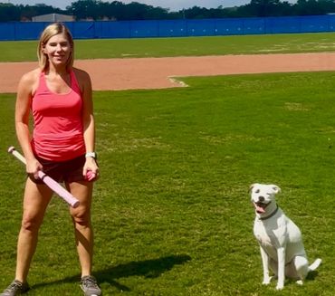 A woman in sportswear holding a bat and ball with a white dog on a baseball field.