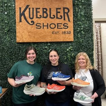 Three women holding colorful Kuebler shoes in front of a green wall with a wooden sign.