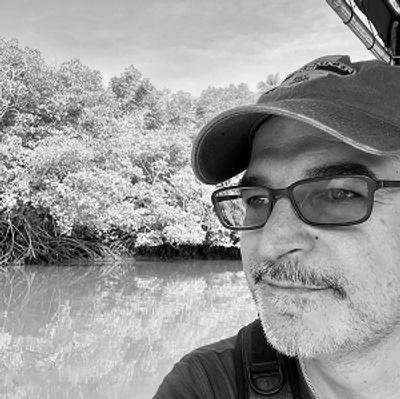Man wearing glasses and cap near a water body with trees in the background.