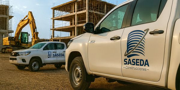 Two white construction trucks with SASEDA logos near an unfinished building and excavator.