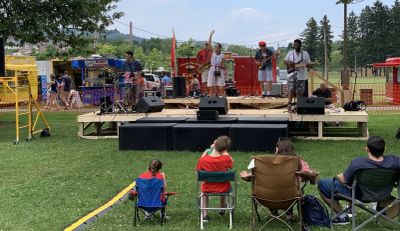 Band on an outdoor festival stage with subwoofers in front, food trucks and a family audience