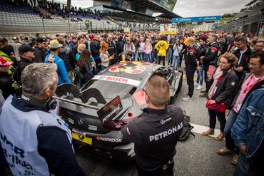 Crowd gathers around a Petronas-sponsored race car during a motorsport event.