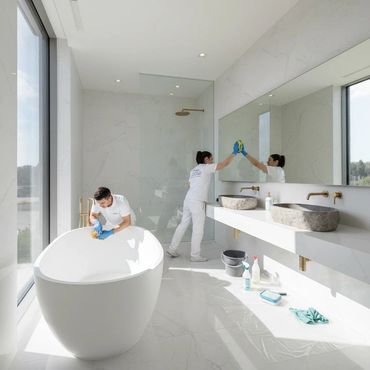 Two cleaners meticulously clean a modern, bright bathroom with large windows.