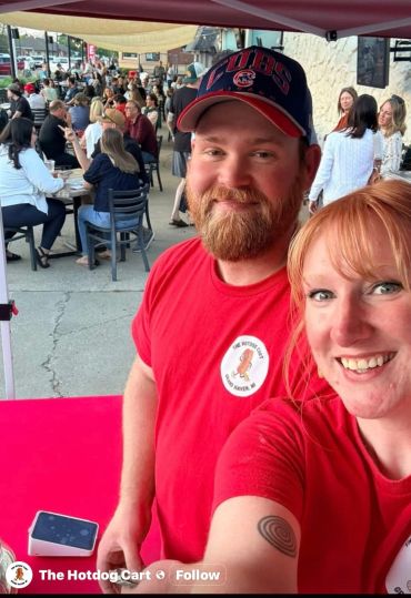 Two smiling vendors in red shirts at a bustling outdoor food stand.