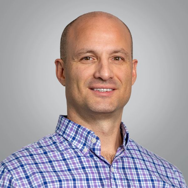 Smiling bald man in a blue and purple plaid shirt against a gray background.
