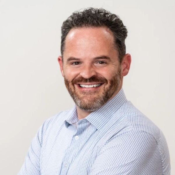 Smiling man with curly hair and beard in a light blue checkered shirt.