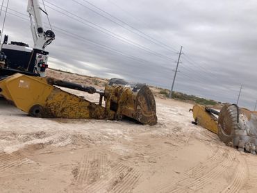 Heavy construction equipment parts lying on a sandy site under a cloudy sky.