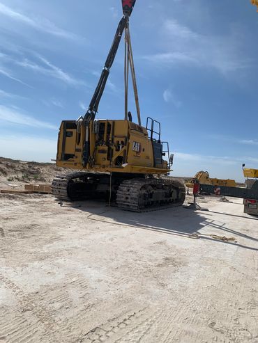 A large yellow CAT excavator being lifted at a construction site under a clear blue sky.