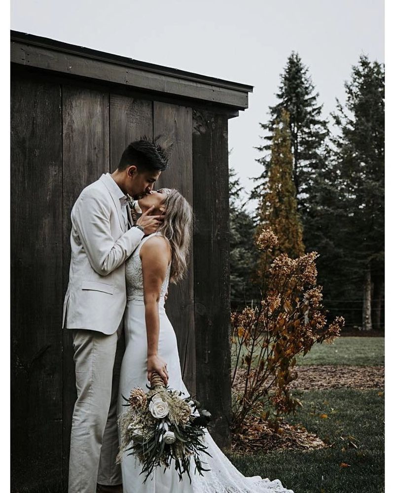 couple up against a barn kissing and holding a white flower with green filler sola wood flower bouqu