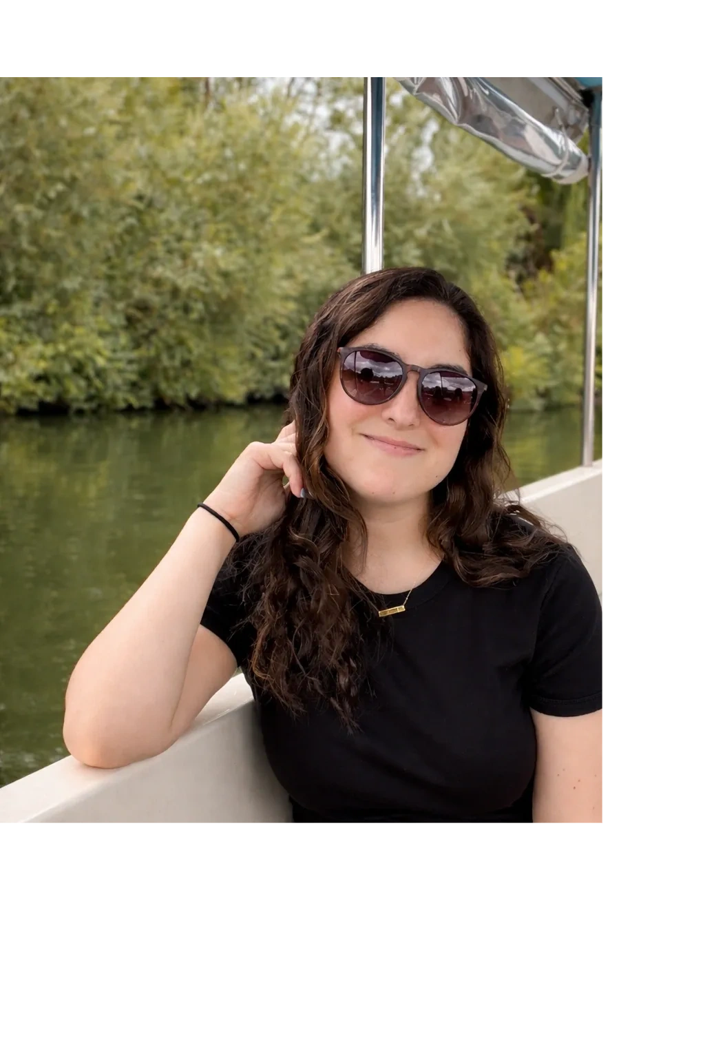 Young woman with sunglasses smiling on a boat near green water and trees.