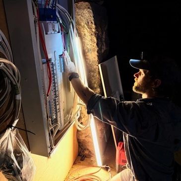 A man works on wiring inside an electrical panel in a dimly lit room.