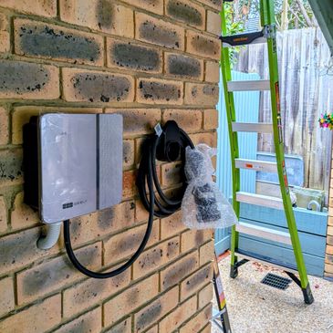 Electric vehicle charging station mounted on a brick wall next to a green ladder.