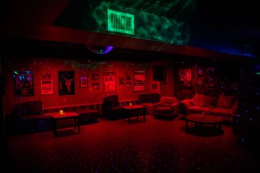 Dimly lit lounge with red and blue lights, cozy seating, and music posters on the walls.