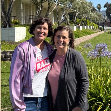 Two women smiling outdoors on a sunny day with greenery and flowers.