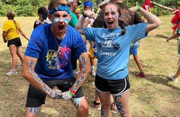 Two enthusiastic teens with blue face paint and arm lettering at an outdoor event.