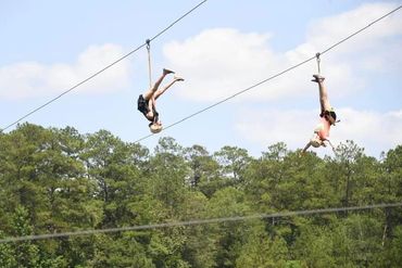 Two people hanging upside down on zip lines over a forested area.