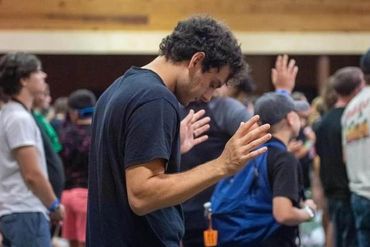 A young man with eyes closed and hands raised among a group of people.