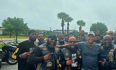 A group of bikers posing with a man in casual sportswear at a gas station.