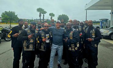A group of bikers posing with a man in casual sportswear at a gas station.