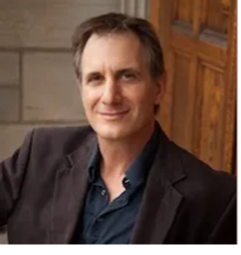 Smiling man in a dark blazer sitting by a wooden door.
