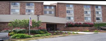 Exterior view of a brick apartment building with landscaped flower beds and an American flag.