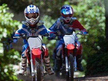 Two kids riding dirt bikes in protective gear on a trail.