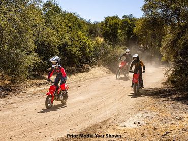 Three kids riding red dirt bikes on a dusty trail surrounded by trees.
