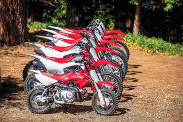 Row of red and white Honda dirt bikes lined up outdoors on a dirt path.