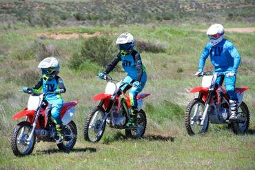 Three riders in blue gear on red dirt bikes riding through grass.